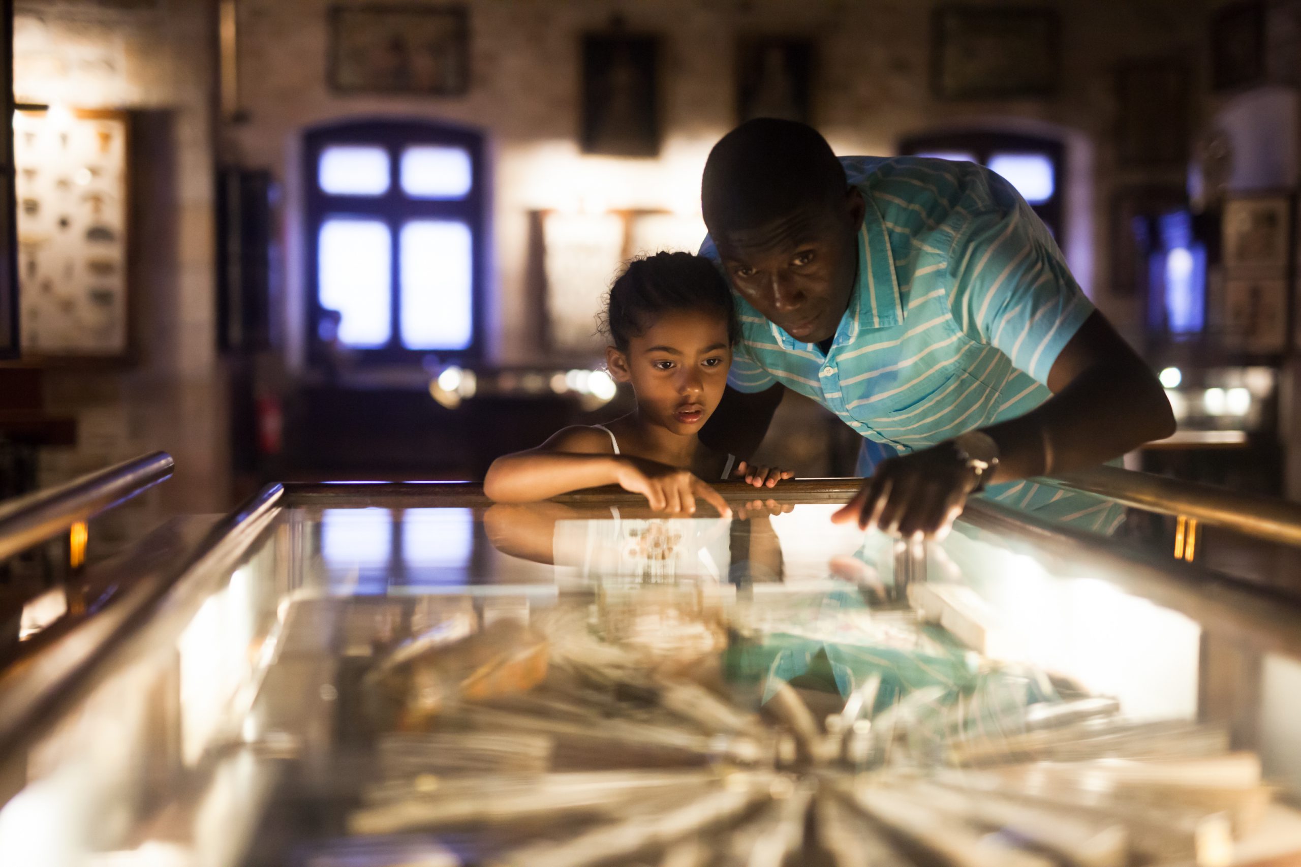 An adult and a child lean over a lit table covered with glass. They are pointing at the exhibit.