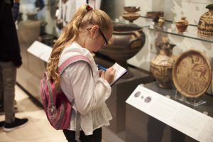 Child in front of cultural artifacts taking notes on paper.