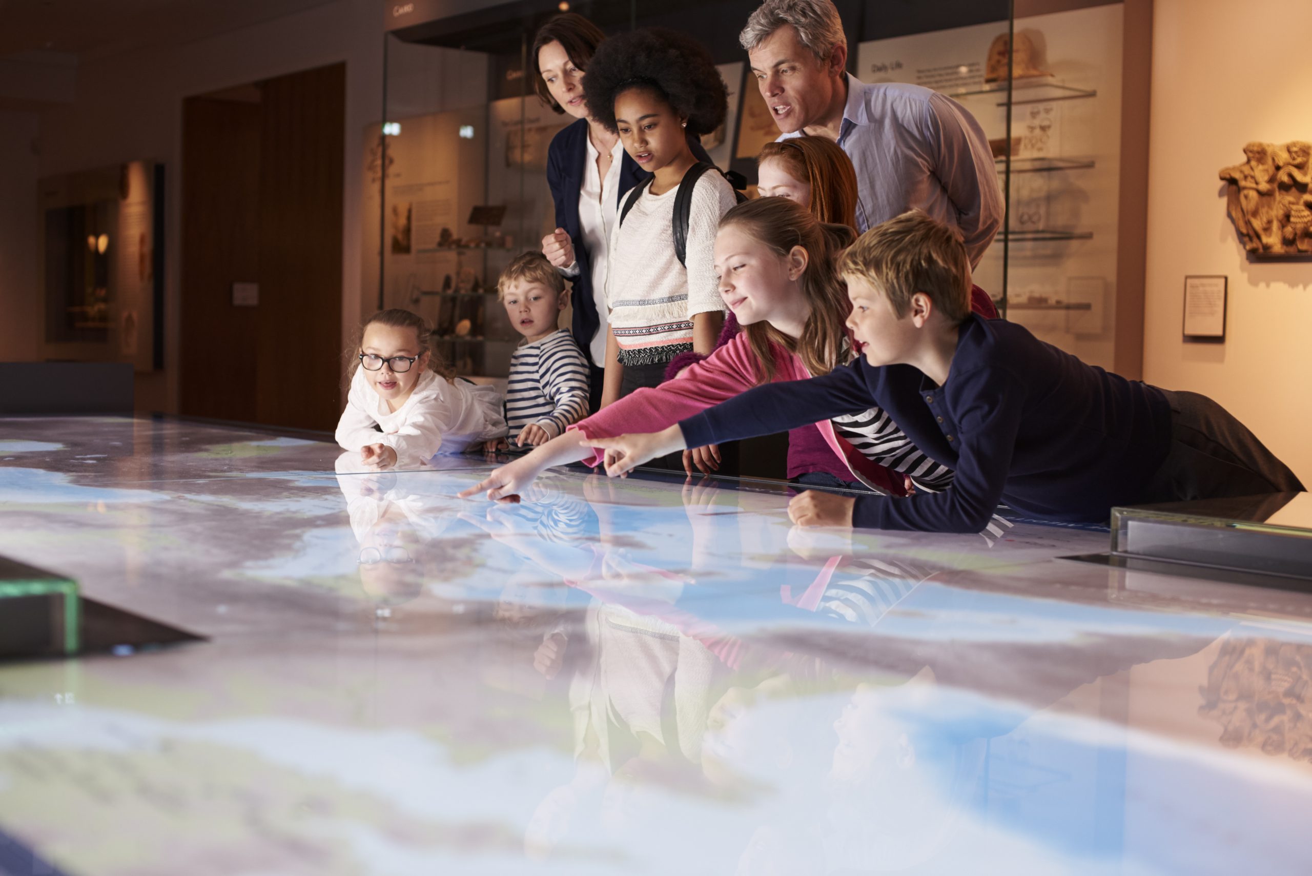 Group of adults and children looking at an interactive table. Three children are leaning over the table and pointing.